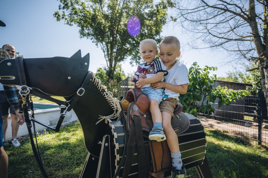 "Zwei Jungen reiten mit Westernsattel auf dem Holzpferd ferdi im Garten