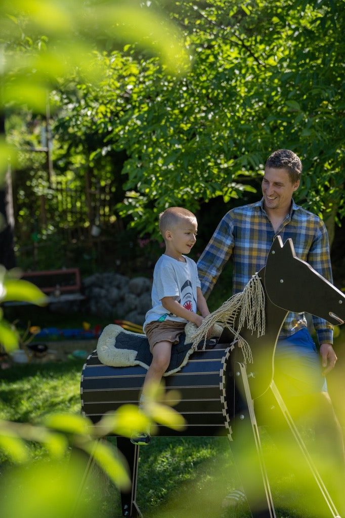 Kind reitet auf stabilem Holzpferd durch Baeume im Garten fotografiert